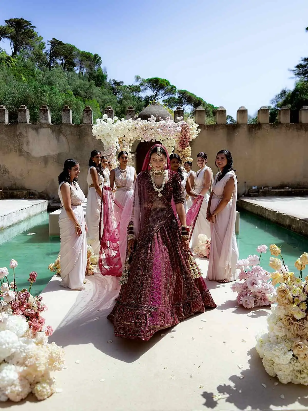 Bride in a red lehenga walking past her bridesmaids beside a courtyard pool